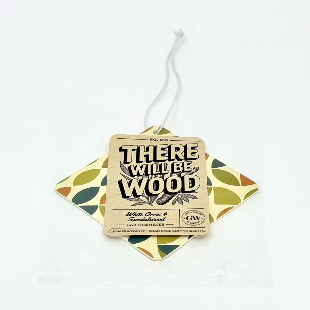 A hanging car freshener tag labeled 'THERE WILL BE WOOD' over a diamond-shaped patterned card in olive green, mustard, and cream, on a plain white background.