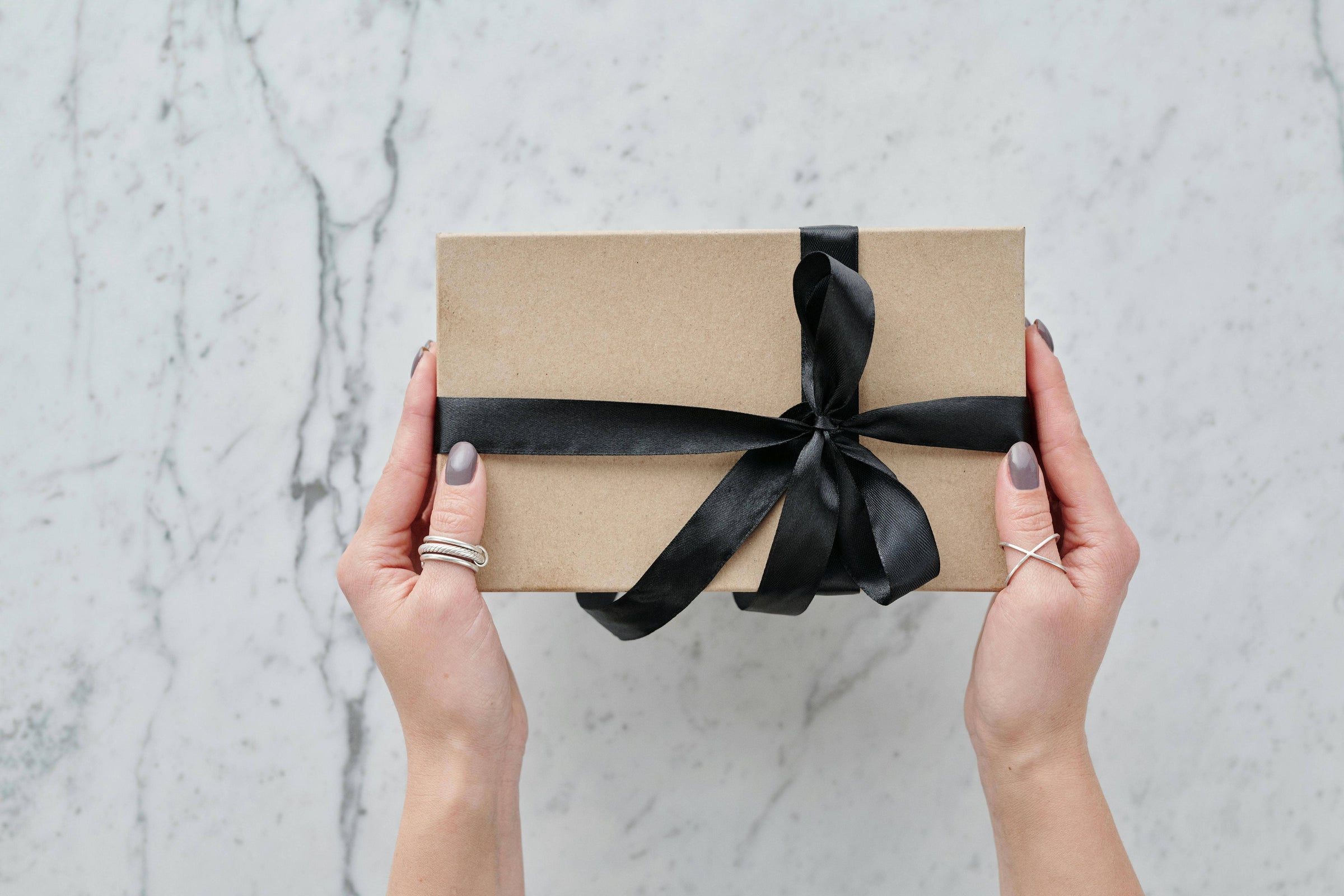 a pair of hands holding a kraft gift box wrapped with black ribbon