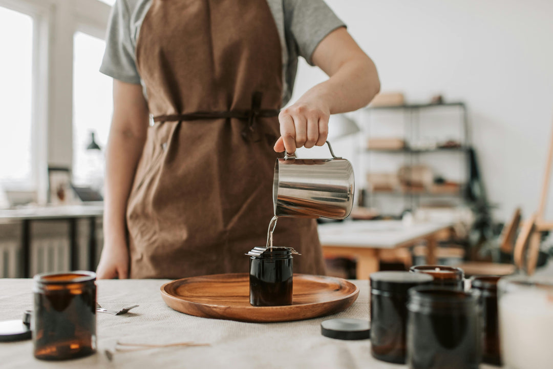 Person in a brown apron pouring wax into a jar on a wooden tray.